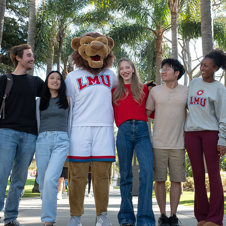 Five students and the LMU lion mascot stand arm in arm on a campus walkway lined with tall palm trees