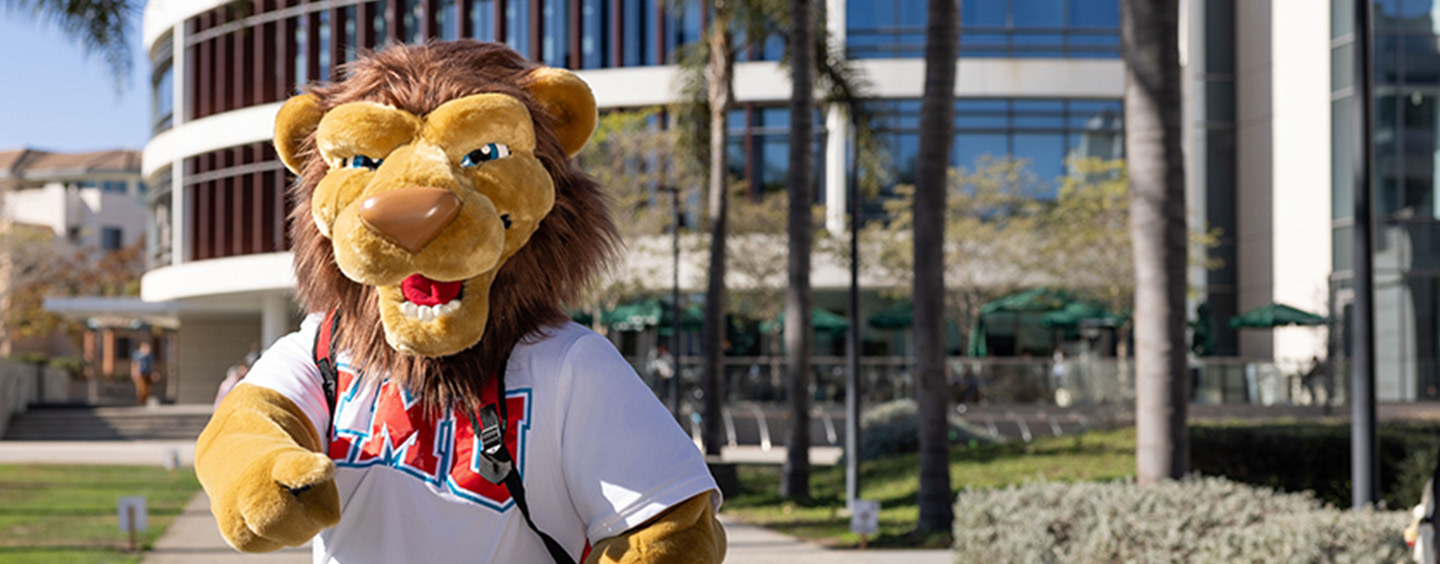 Iggy, the mascot, stands outside in a white LMU shirt with the LMU library.
