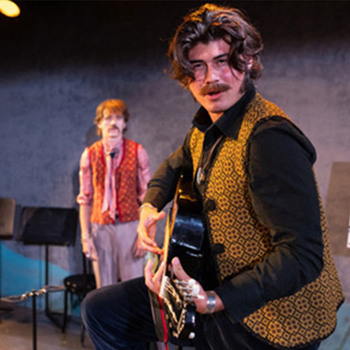 A theatrical performance scene featuring a person in the foreground playing a black guitar while wearing a patterned gold-and-black vest over dark clothing