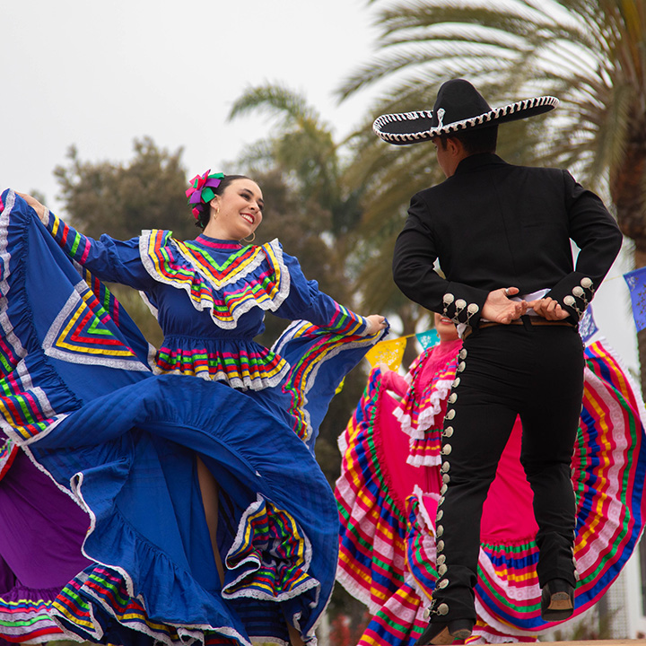 Traditional folklórico dancers perform outdoors on a campus lawn, wearing brightly colored dresses with multicolored ribbon trim and a black charro-style outfit with a wide-brimmed sombrero