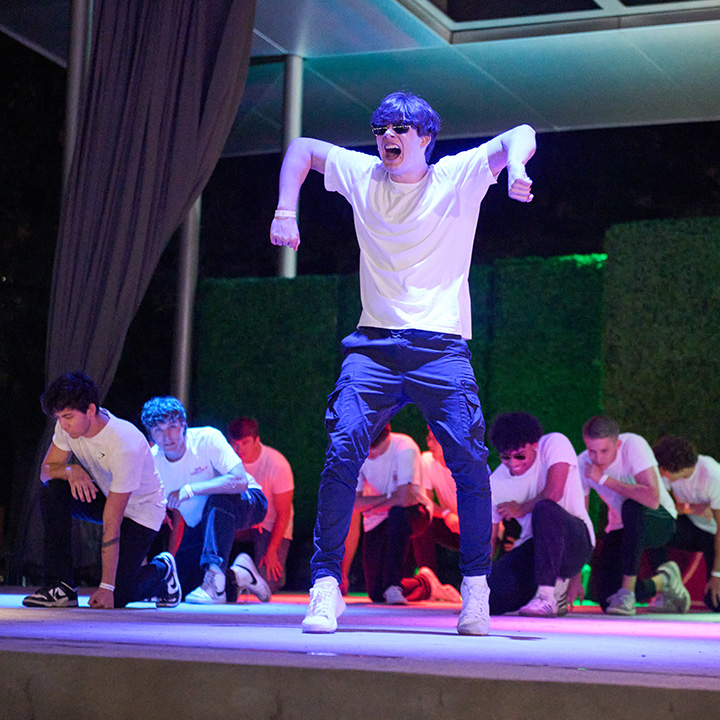 A group performs a choreographed dance on an outdoor stage at night. One performer in a white T-shirt, dark pants, and sunglasses stands at center stage in a dynamic pose, while several other performers kneel or crouch behind in coordinated positions