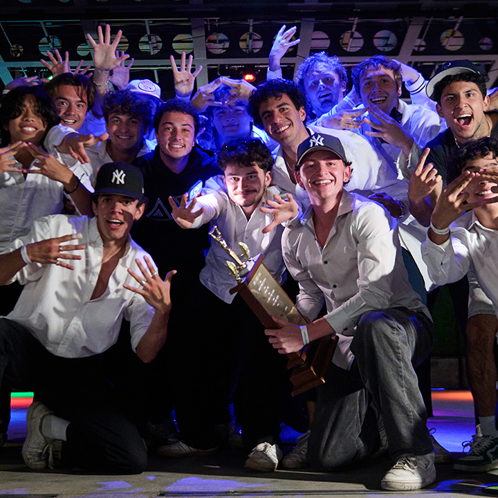 Male students gather cheering and holding a trophy in white sweatshirts on a stage.