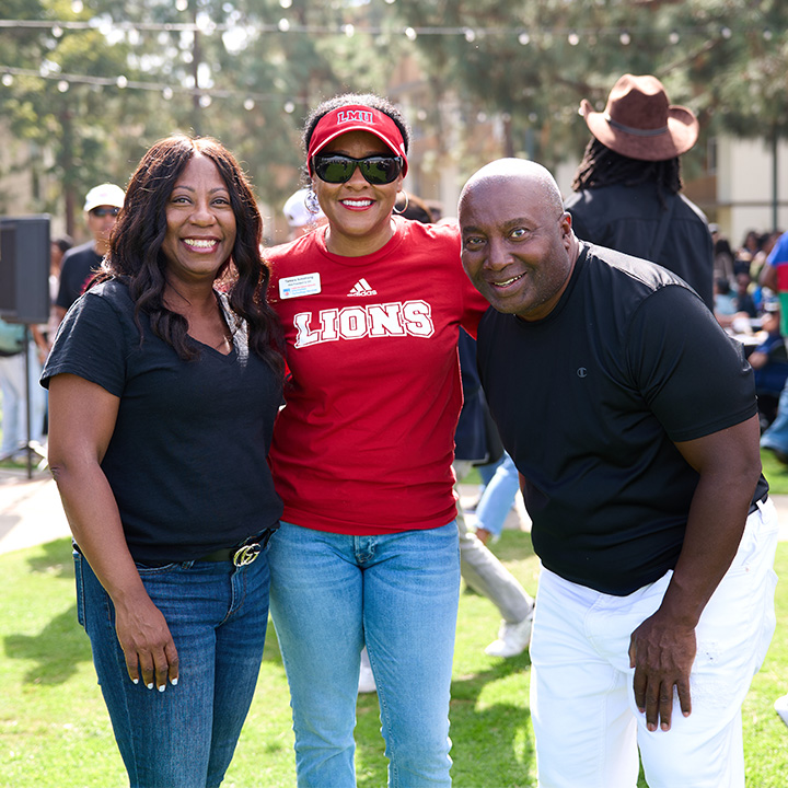 Three people stand together outdoors on a campus lawn beside a large tree during a daytime event