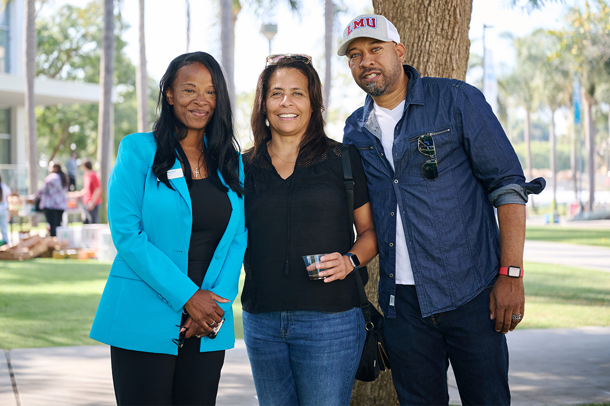 Three people stand together outdoors on a campus lawn beside a large tree during a daytime event