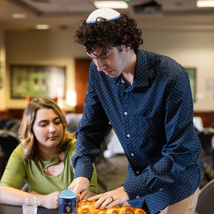 A person wearing a white kippah stands at a table cutting a braided loaf of challah bread during an indoor gathering. Another person seated nearby watches the ceremony.