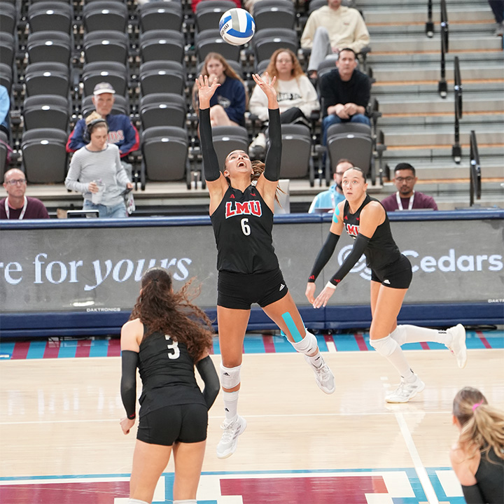An LMU volleyball player wearing jersey number 6 jumps with both hands raised to set a volleyball during a match. Teammates in black uniforms surround the player on the court, while spectators sit in the arena seats in the background. Visible LMU branding appears on the uniform and around the court.