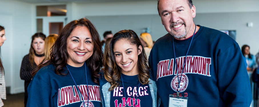 Mother, father and female daughter standing together inside wearing blue LMU shirts